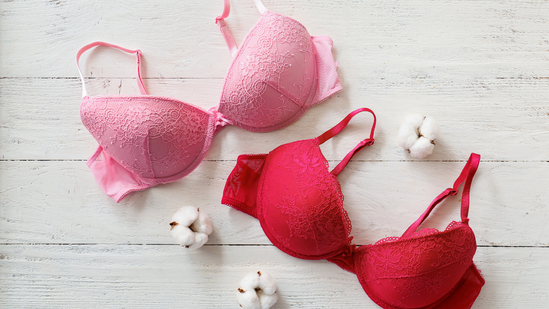 Two lace bras in shades of pink and red laid out on a white wooden surface, surrounded by cotton flowers