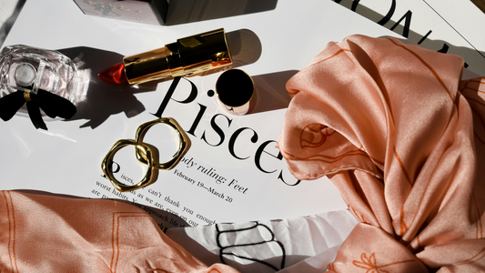 Flat lay of feminine essentials including a peach silk scarf, gold rings, red lipstick, and a perfume bottle