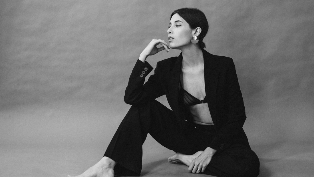 A confident woman poses barefoot in a tailored black suit with a sheer black bra visibly styled under the blazer