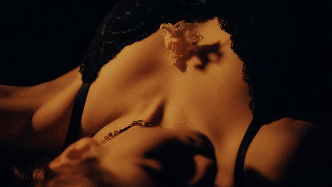 Close-up of a woman wearing a black lace bra, softly lit in warm golden tones, with a delicate flower placed on her chest